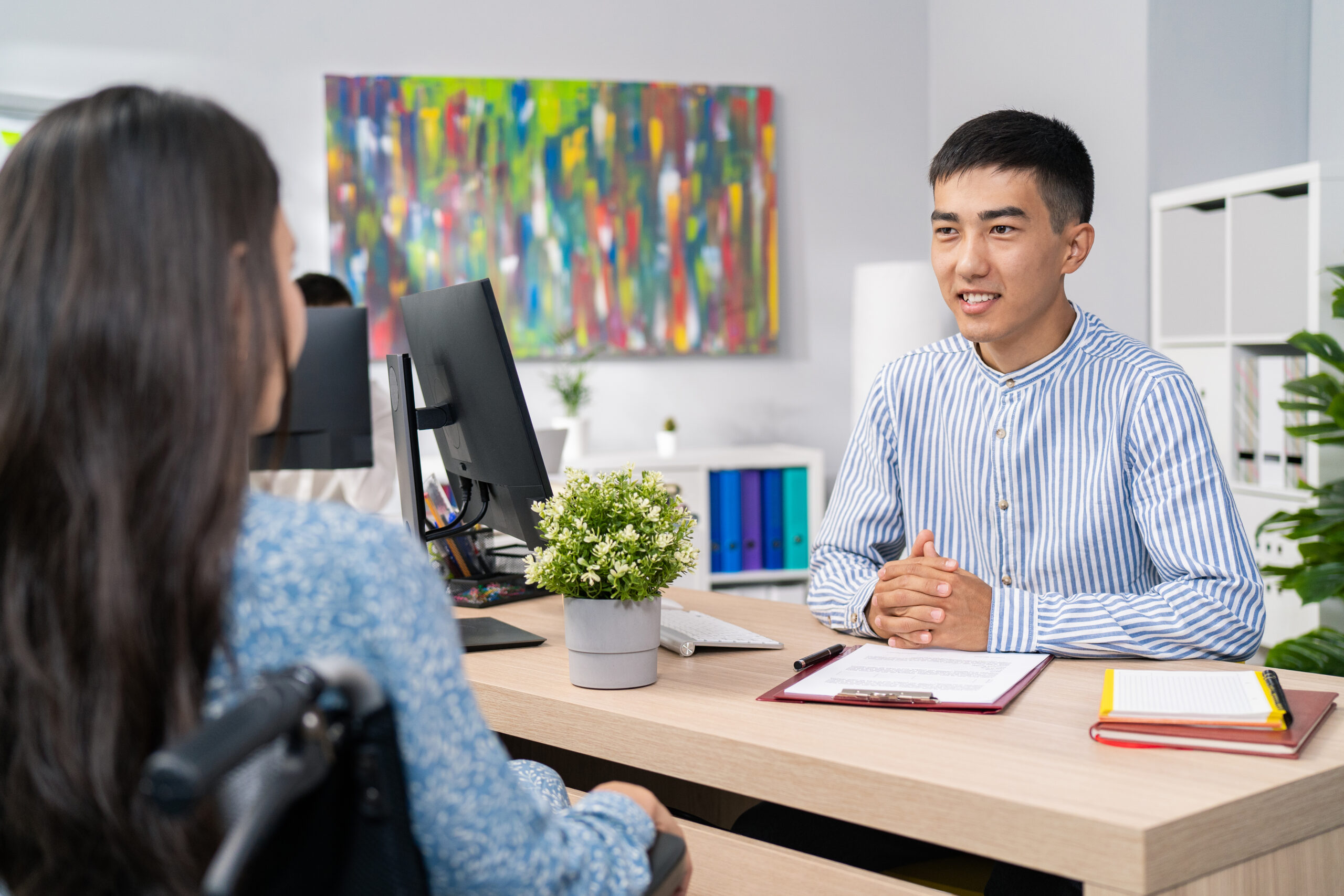 Job interview woman with disability use wheelchair, stern male boss asks questions to girl in dress, they sit in company office on two sides of desk guy listens carefully to answer