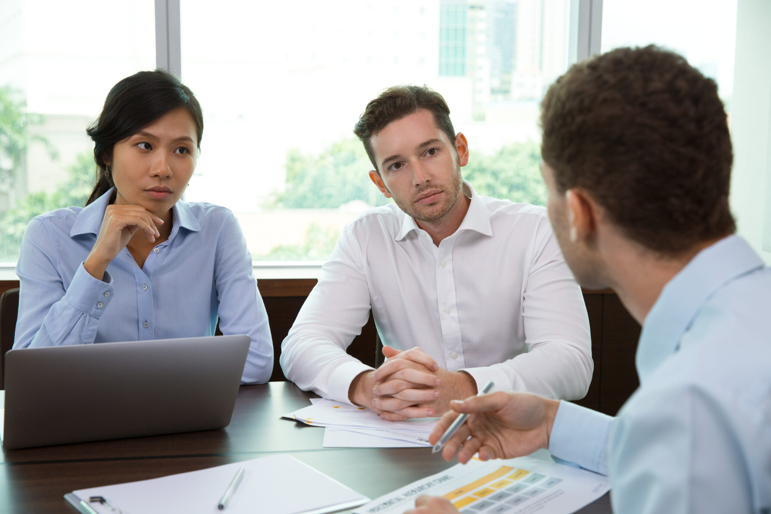 Young businessman and businesswoman sitting at desk with office window in background and listening to their male colleague sitting back to camera